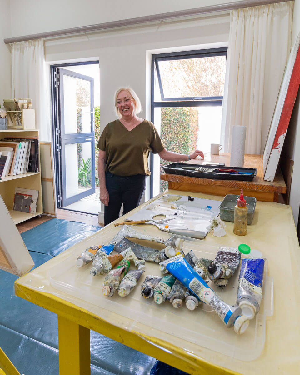 Janet Dirksen smiling in her art studio, paints and tools in front of her.